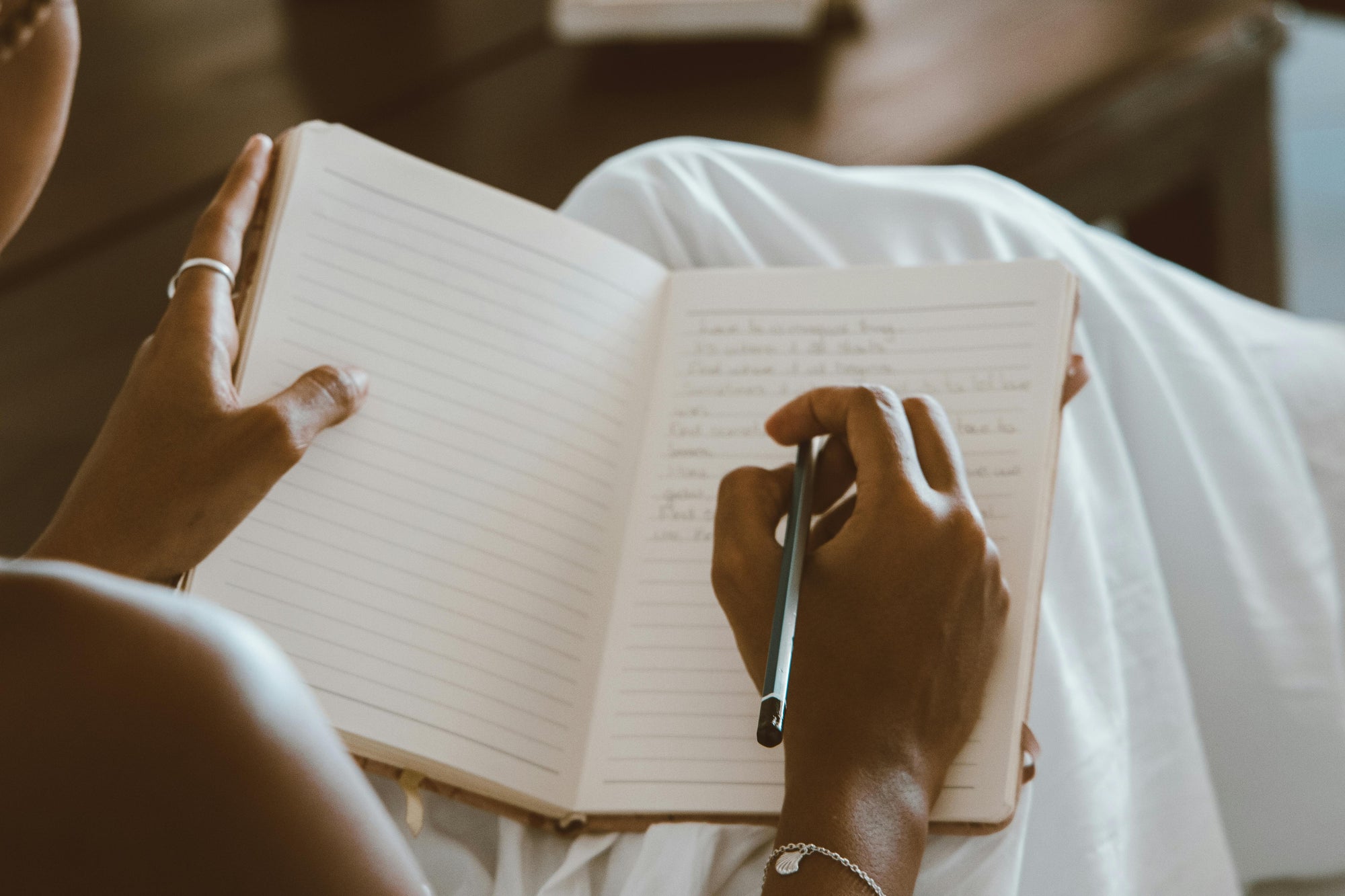 Person writing in a lined journal while sitting cross-legged, holding a pen in one hand and resting the notebook on their lap